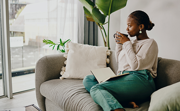 Young woman sitting on a couch, holding a coffee and looking out a window.